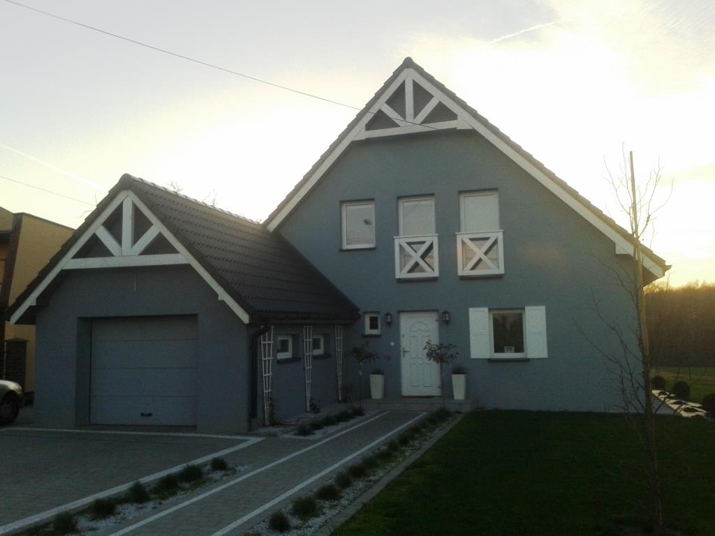 Gotowy dom jednorodzinny z garażem, pomalowany na szaro, z białymi detalami architektonicznymi i zagospodarowanym terenem wokół.