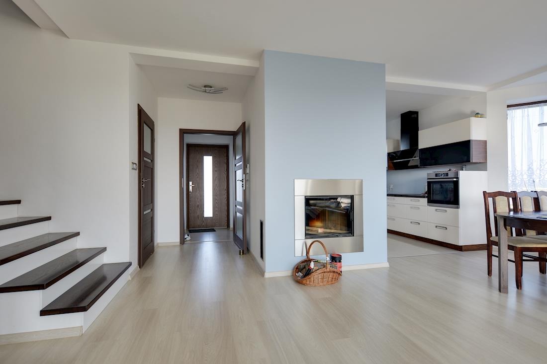 Jasne wnętrze domu z drewnianymi schodami, drzwiami wejściowymi, kominkiem obudowanym w stal oraz widoczną kuchnią w tle.