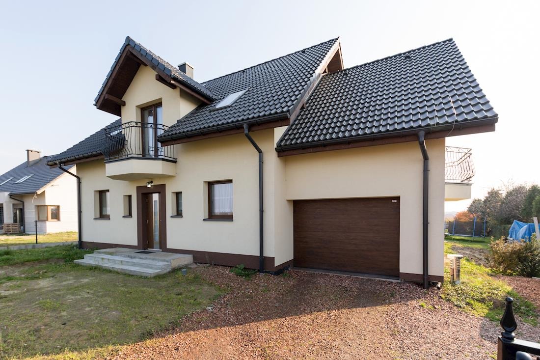 Gotowy, dwukondygnacyjny dom z garażem i balkonem, wykończony w jasnych kolorach z ciemną dachówką, widok z zewnątrz na tle błękitnego nieba.