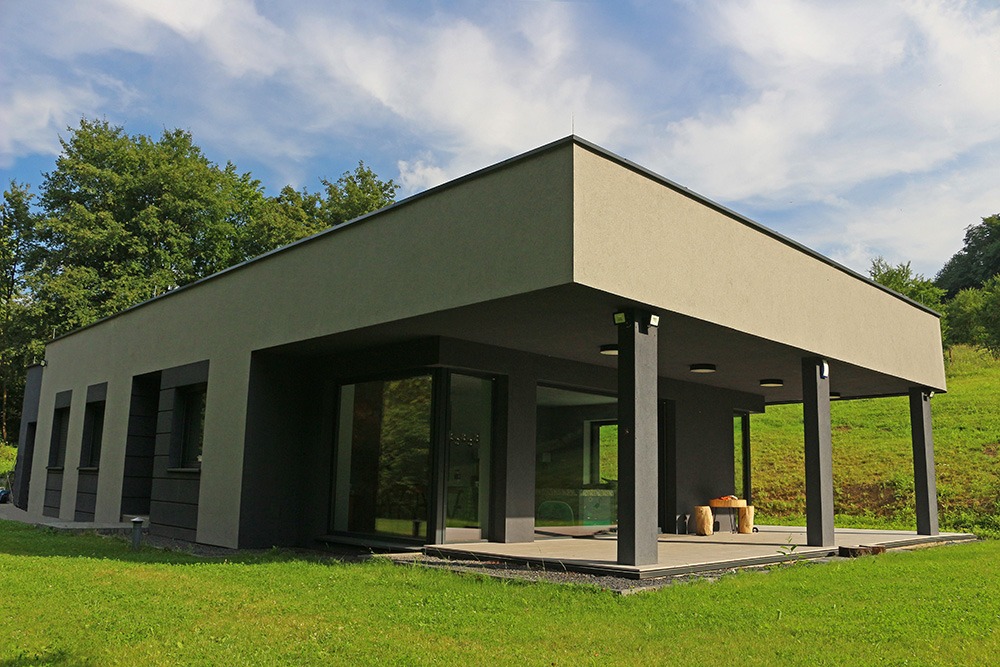 Nowoczesny dom parterowy z płaskim dachem, wykończony w odcieniach szarości i beżu, z dużymi przeszkleniami i zadaszonym tarasem, otoczony zielonym trawnikiem na tle zalesionego wzgórza.