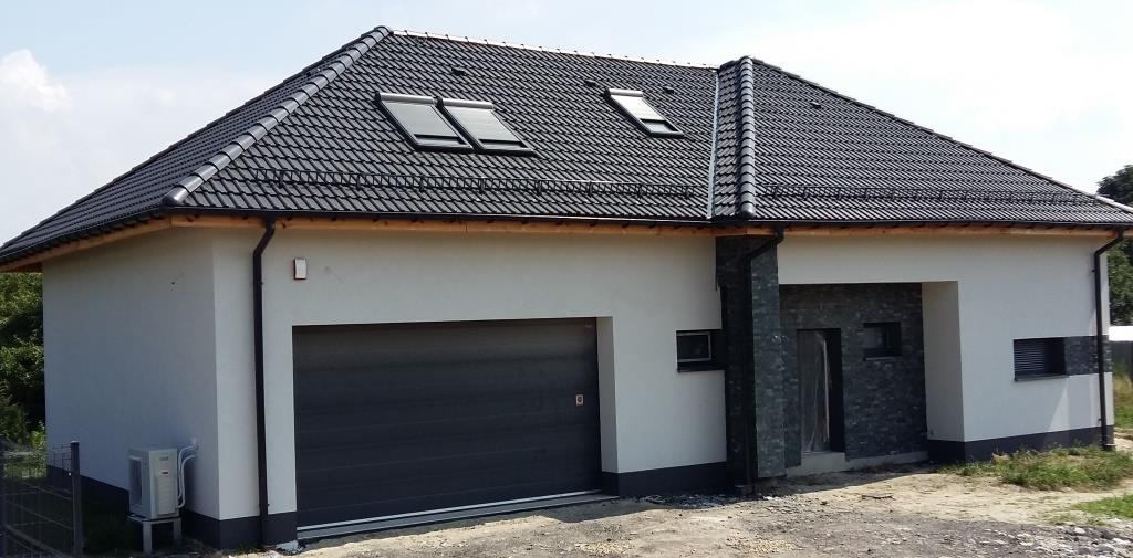 Nowoczesny parterowy budynek z garażem i czarną dachówką, widoczne okna dachowe, elewacja w jasnym odcieniu szarości, fragment dekoracyjnej ściany z kamienia.
