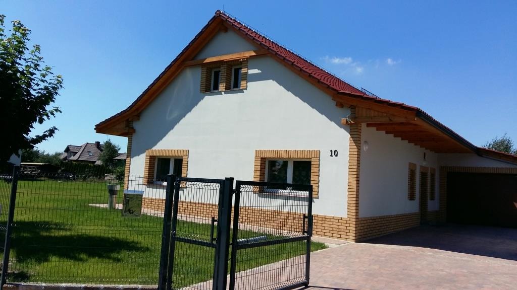 Nowo wybudowany dom jednorodzinny z czerwonym dachem i elewacją w kolorze złamanej bieli, z numerem '10' na ścianie, otoczony zielonym trawnikiem i czarnym ogrodzeniem z metalowej siatki.