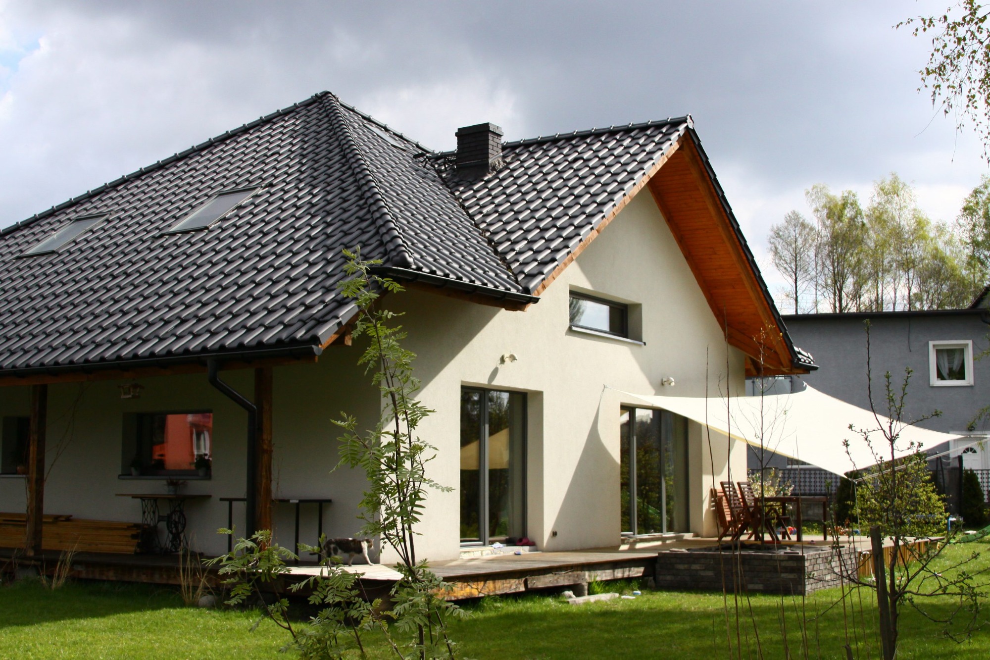 Nowoczesny dom z ciemną dachówką i dużymi oknami tarasowymi, z zadaszonym tarasem i markizą przeciwsłoneczną w ogrodzie.