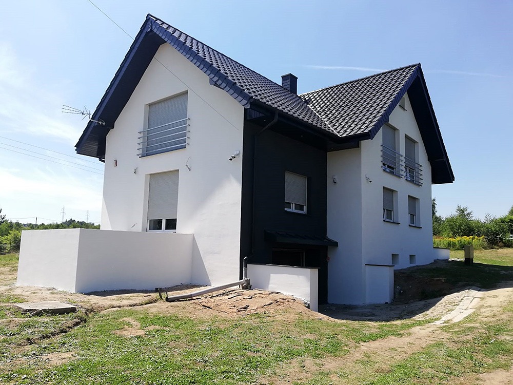 Nowoczesny dom murowany z dwuspadowym dachem i białymi ścianami, z ciemnoszarym fragmentem elewacji oraz widocznymi roletami zewnętrznymi i anteną satelitarną na dachu. Teren wokół domu w trakcie...