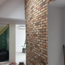 Ściana z cegły dekoracyjnej w trakcie prac wykończeniowych, widoczna instalacja elektryczna, wiadro i drabina w tle, surowy stan pomieszczenia.