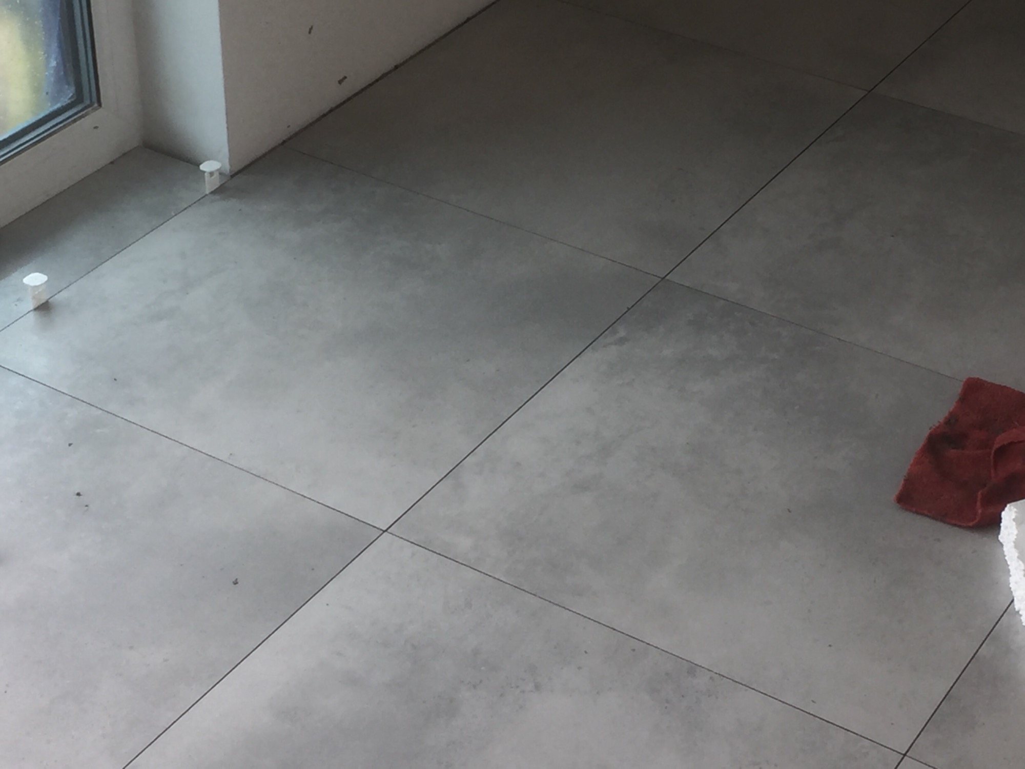 Szare płytki podłogowe ułożone w kwadraty, widoczne krzyżyki dystansowe przy ścianie i czerwona ścierka w rogu.