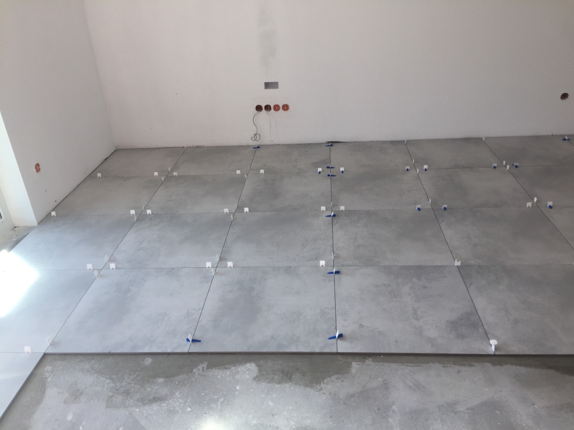 Szare płytki podłogowe ułożone w rzędach, z użyciem białych i niebieskich krzyżyków dystansowych, widoczne gniazdka elektryczne w ścianie, podłoże przed płytkami jeszcze nie wykończone.