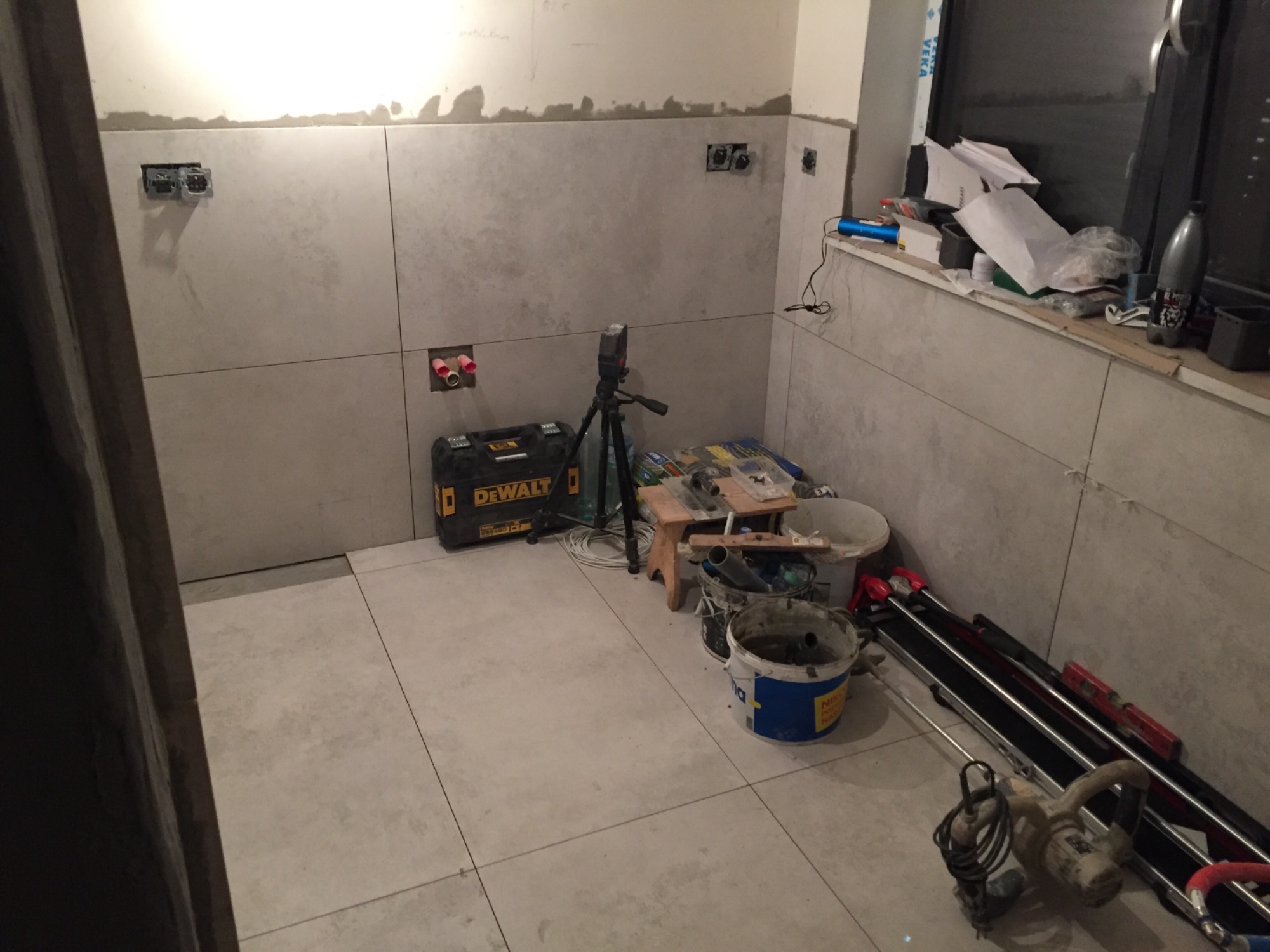 Wnętrze łazienki w trakcie układania płytek, widoczne narzędzia: laser krzyżowy na statywie, wiadro z zaprawą, przecinarka do płytek, skrzynka narzędziowa DeWalt, puszki elektryczne przygotowane...