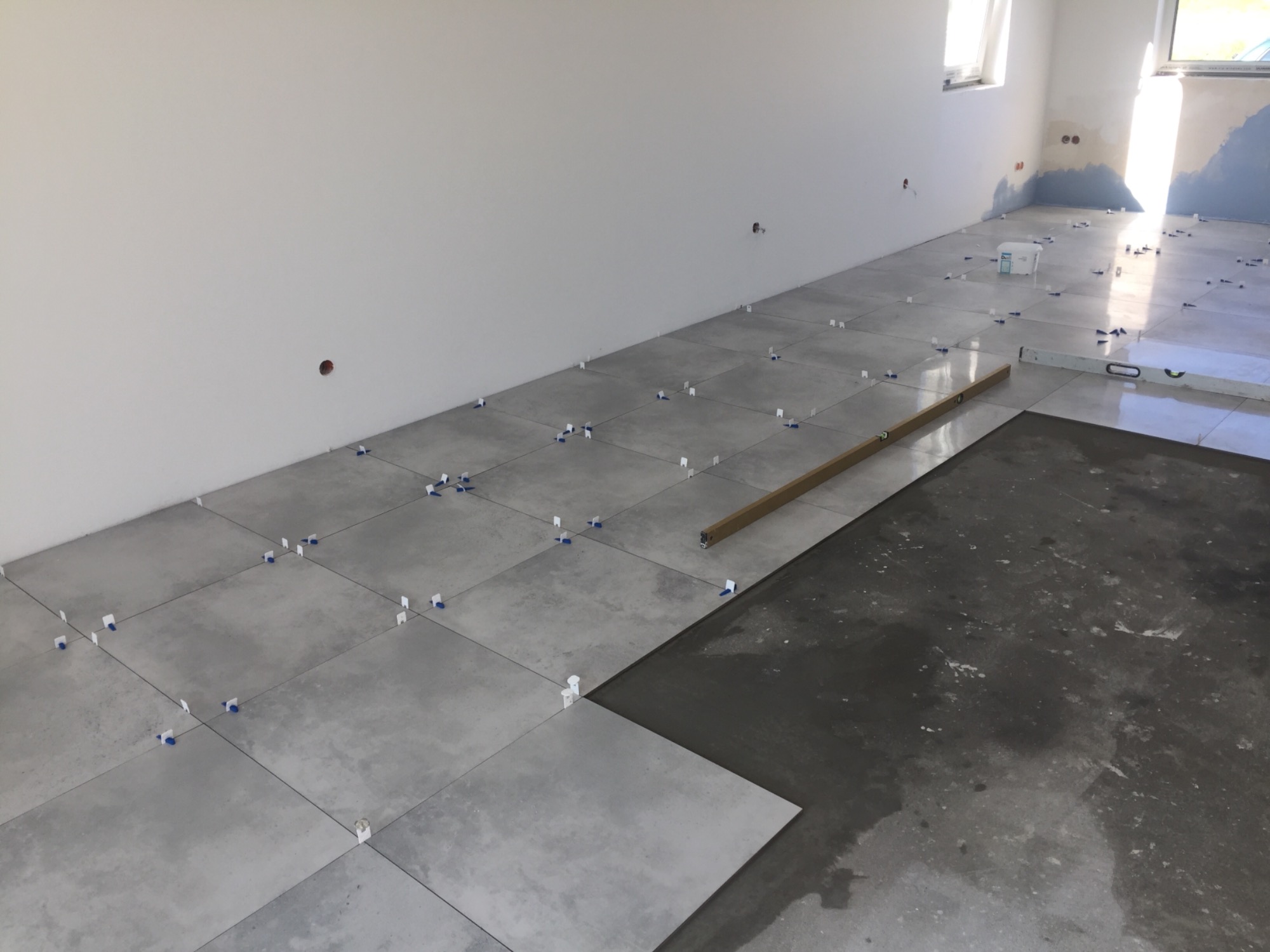 Proces układania szarych płytek podłogowych z użyciem poziomicy i niebieskich krzyżyków dystansowych, widoczny fragment podłogi przed położeniem płytek.