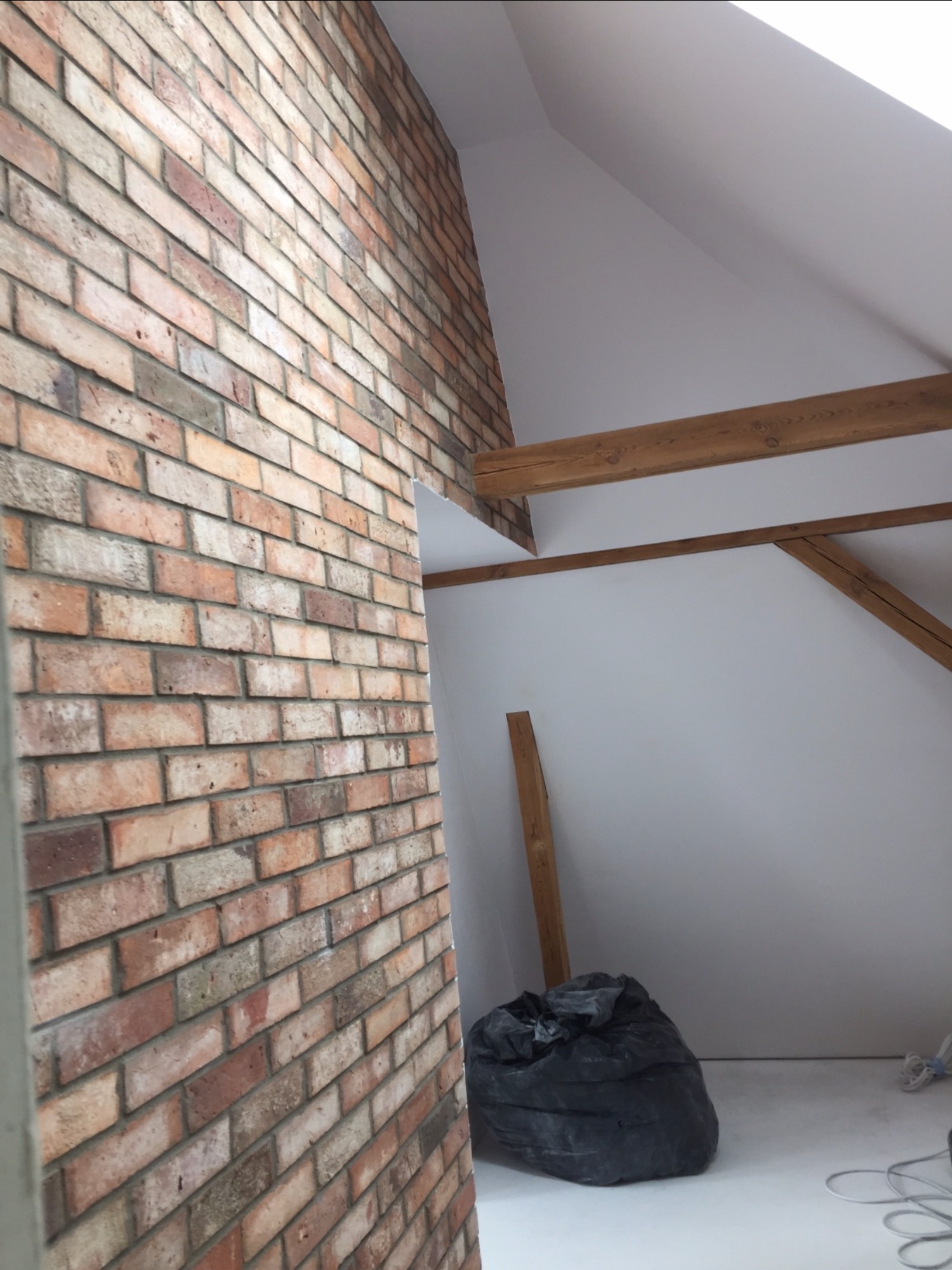 Ściana z cegły dekoracyjnej w poddaszowym pokoju z drewnianymi belkami stropowymi, worek na śmieci w rogu pomieszczenia.