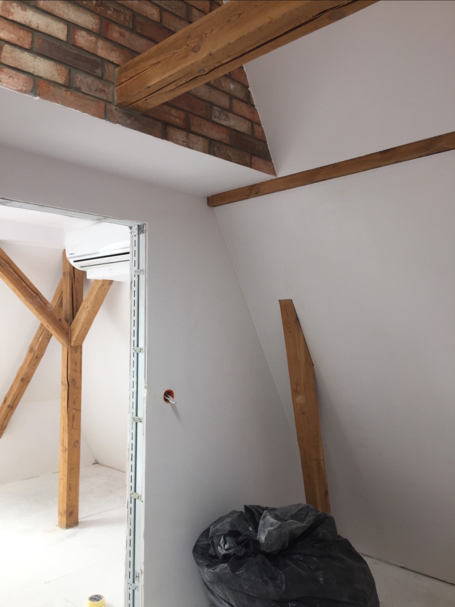 Poddasze w trakcie prac wykończeniowych: białe ściany, drewniane belki stropowe, fragment muru z cegły, klimatyzator, czarny worek na śmieci w rogu.