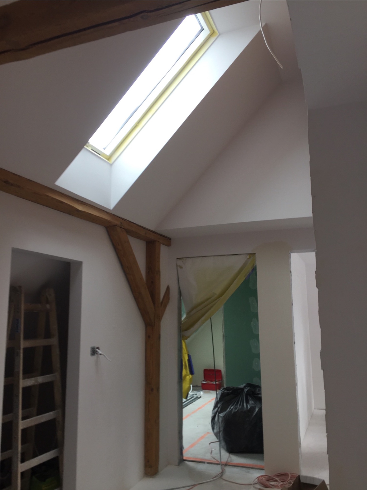 Wnętrze poddasza w trakcie prac wykończeniowych: widoczne drewniane elementy konstrukcyjne, okno dachowe, ściany przygotowane pod malowanie, drabina i worki z odpadami budowlanymi.