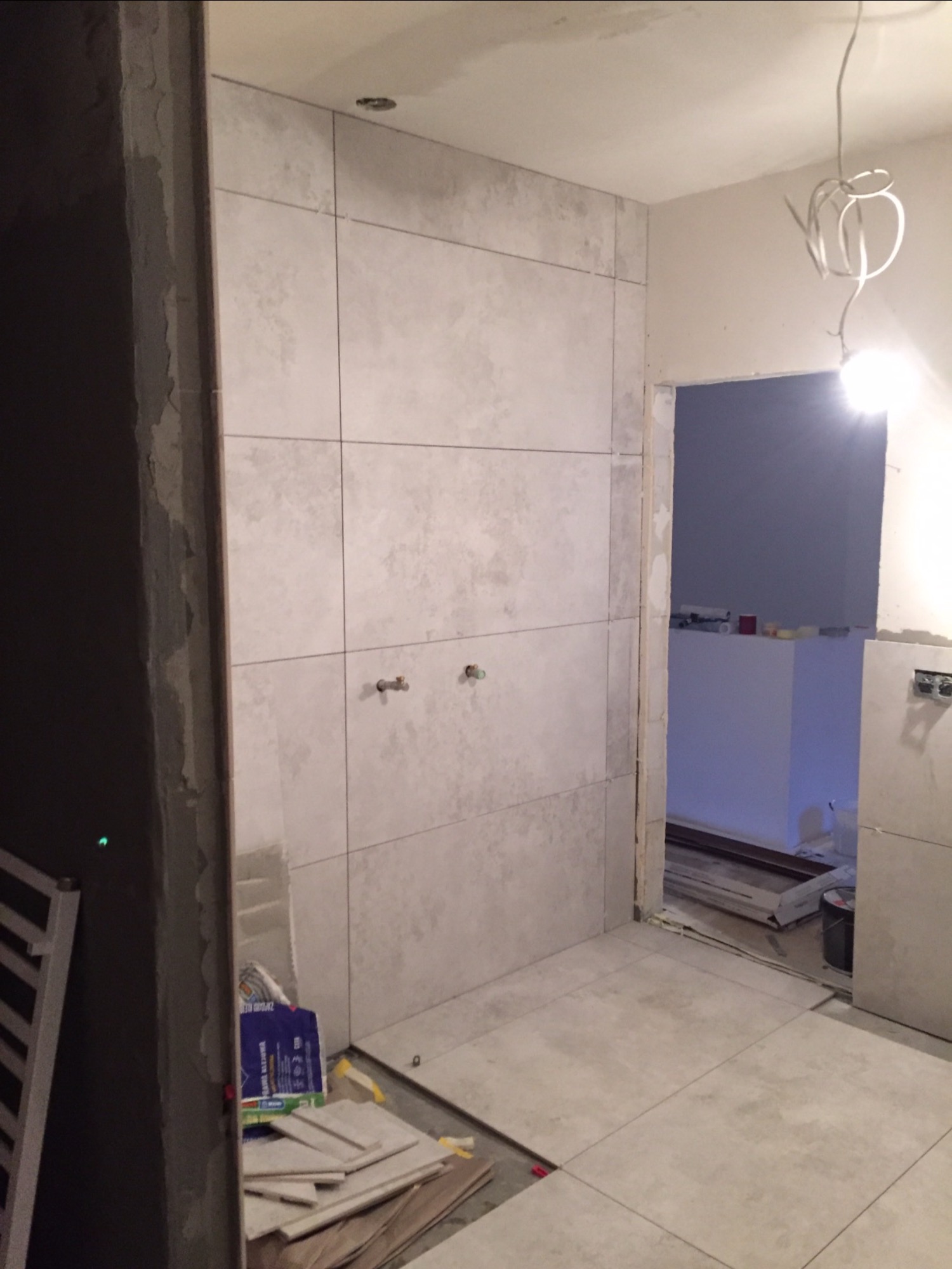 Wnętrze łazienki w trakcie prac, widoczne płytki na ścianach i podłodze, wystające rury instalacji wodnej, oraz materiały budowlane w rogu pomieszczenia.