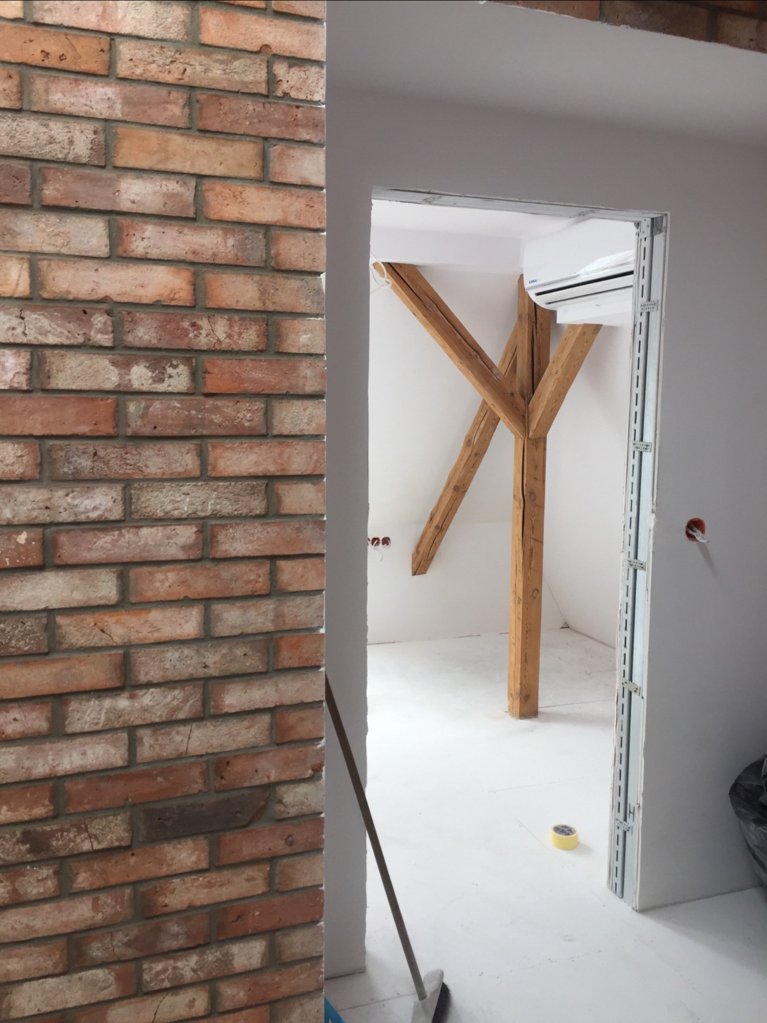 Ściana z cegły klinkierowej w surowym wnętrzu z drewnianą konstrukcją nośną i klimatyzatorem, widok przez otwór drzwiowy w trakcie remontu.