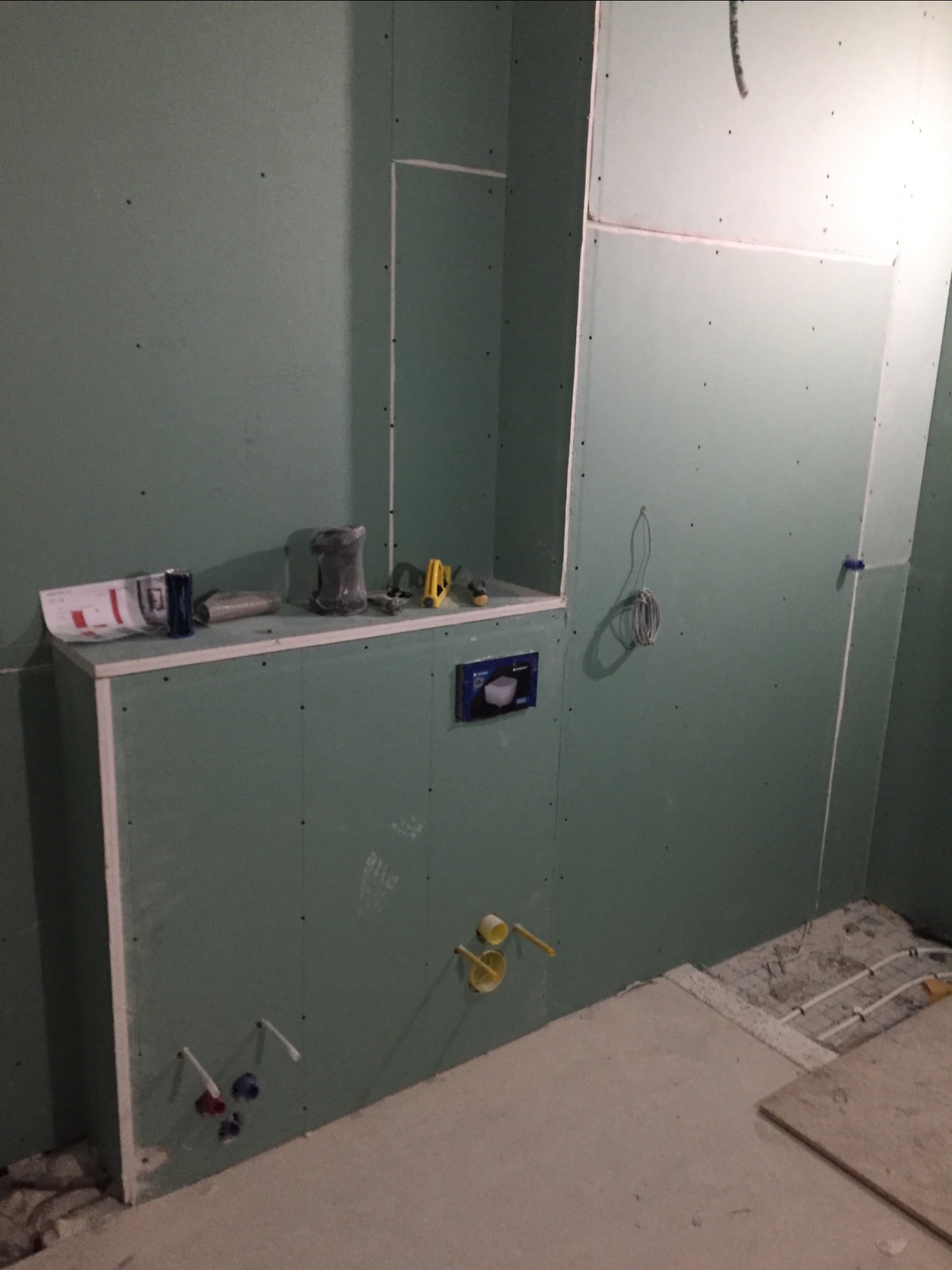 Wnętrze łazienki w trakcie prac wykończeniowych: ściany obłożone płytami gipsowo-kartonowymi, widoczne instalacje wodno-kanalizacyjne, puszka podtynkowego WC, narzędzia na półce, fragment...