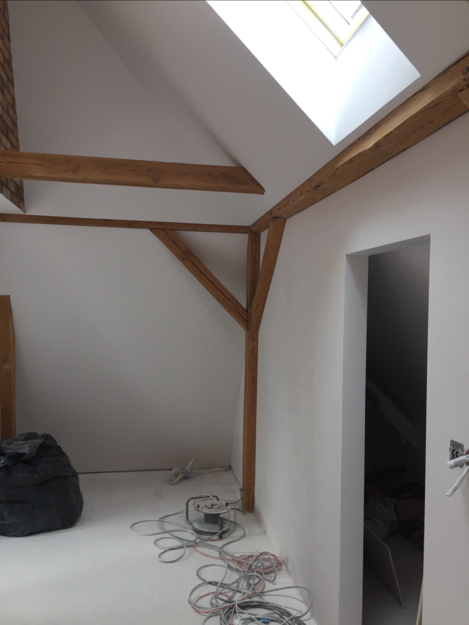 Poddasze po remoncie z widocznymi drewnianymi belkami konstrukcyjnymi, oknem dachowym i przygotowanym podłożem pod posadzkę, z widocznym workiem na śmieci i szlifierką do podłóg.
