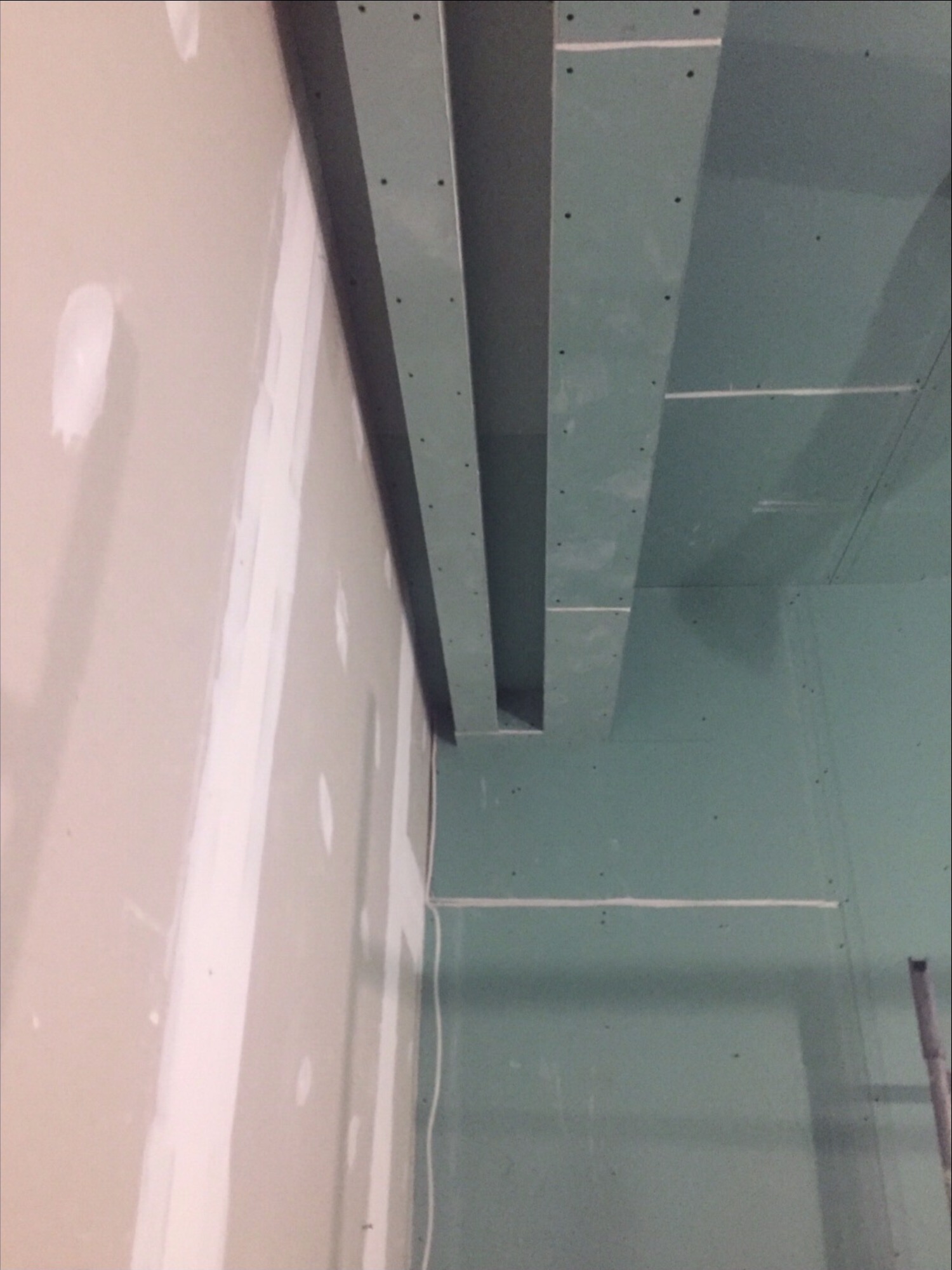 Ściana z płyt gipsowo-kartonowych w trakcie montażu, widoczne łączenia płyt, elementy konstrukcyjne sufitu podwieszanego i przewody elektryczne.