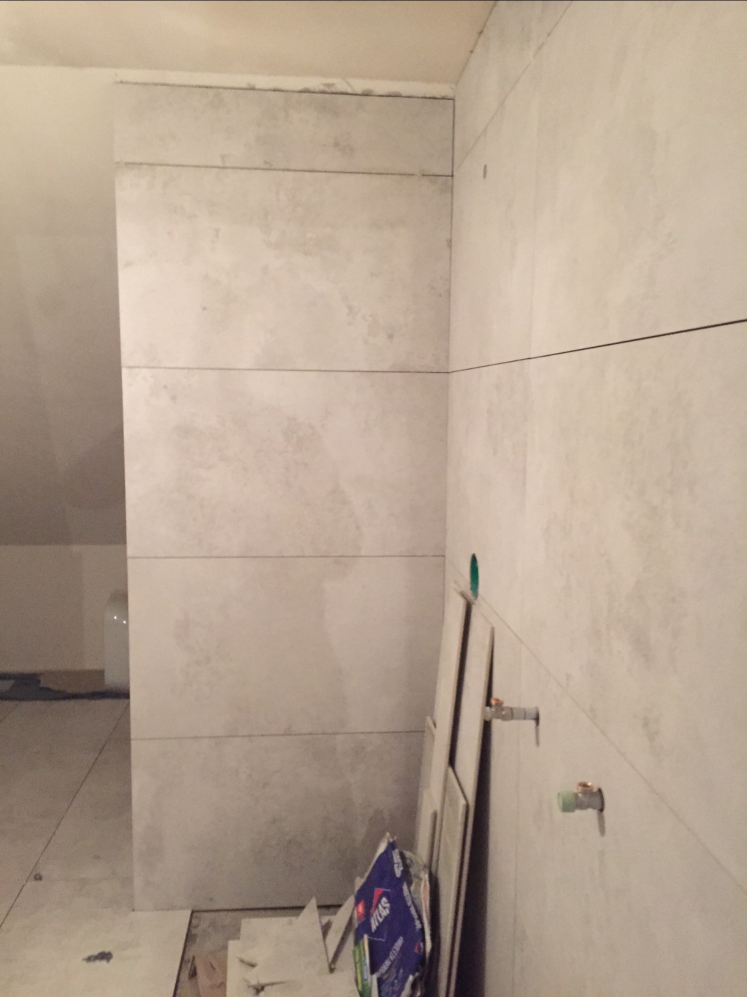 Ściana w łazience w trakcie wykańczania, widoczne płytki o wzorze imitującym kamień, wystające przyłącza wodne, paczka kleju do płytek i nieułożone płytki na podłodze.