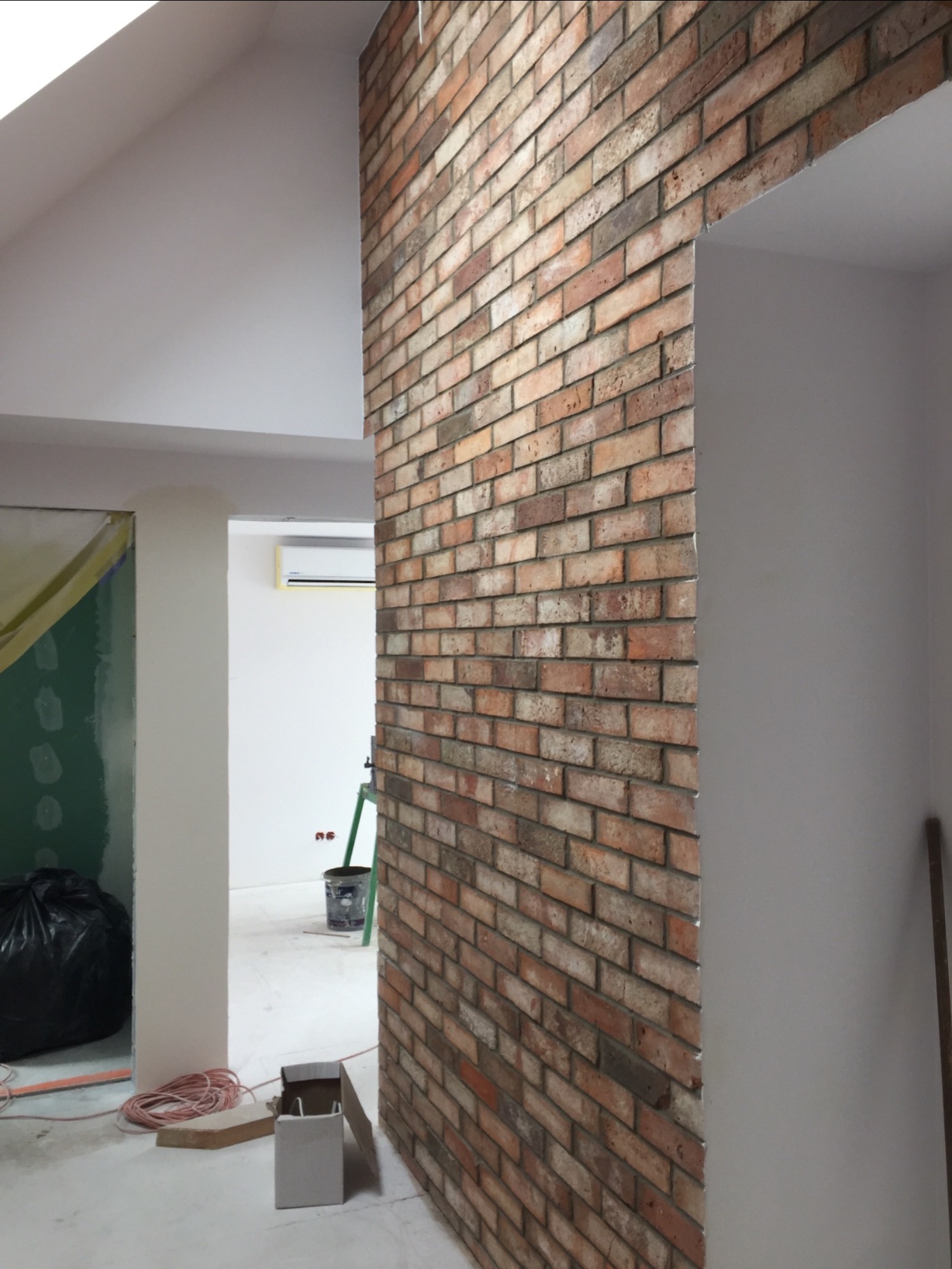 Ściana z cegły dekoracyjnej w trakcie prac wykończeniowych, widoczna instalacja elektryczna, wiadro i drabina w tle, surowy stan pomieszczenia.