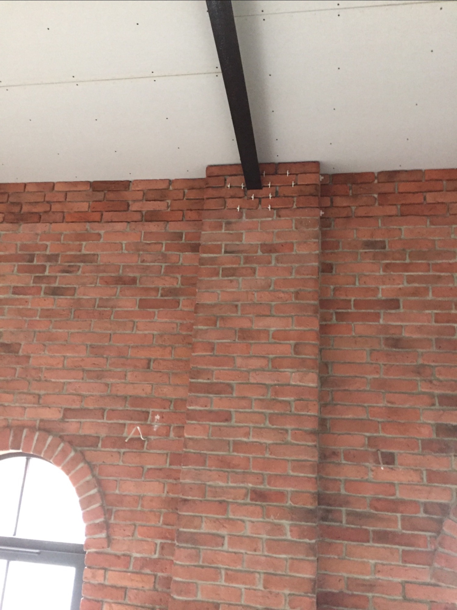 Czerwona ściana z cegły z fragmentem okna łukowego, biały sufit z widocznymi wkrętami i czarną belką, widoczne krzyżyki dystansowe na górze ściany.