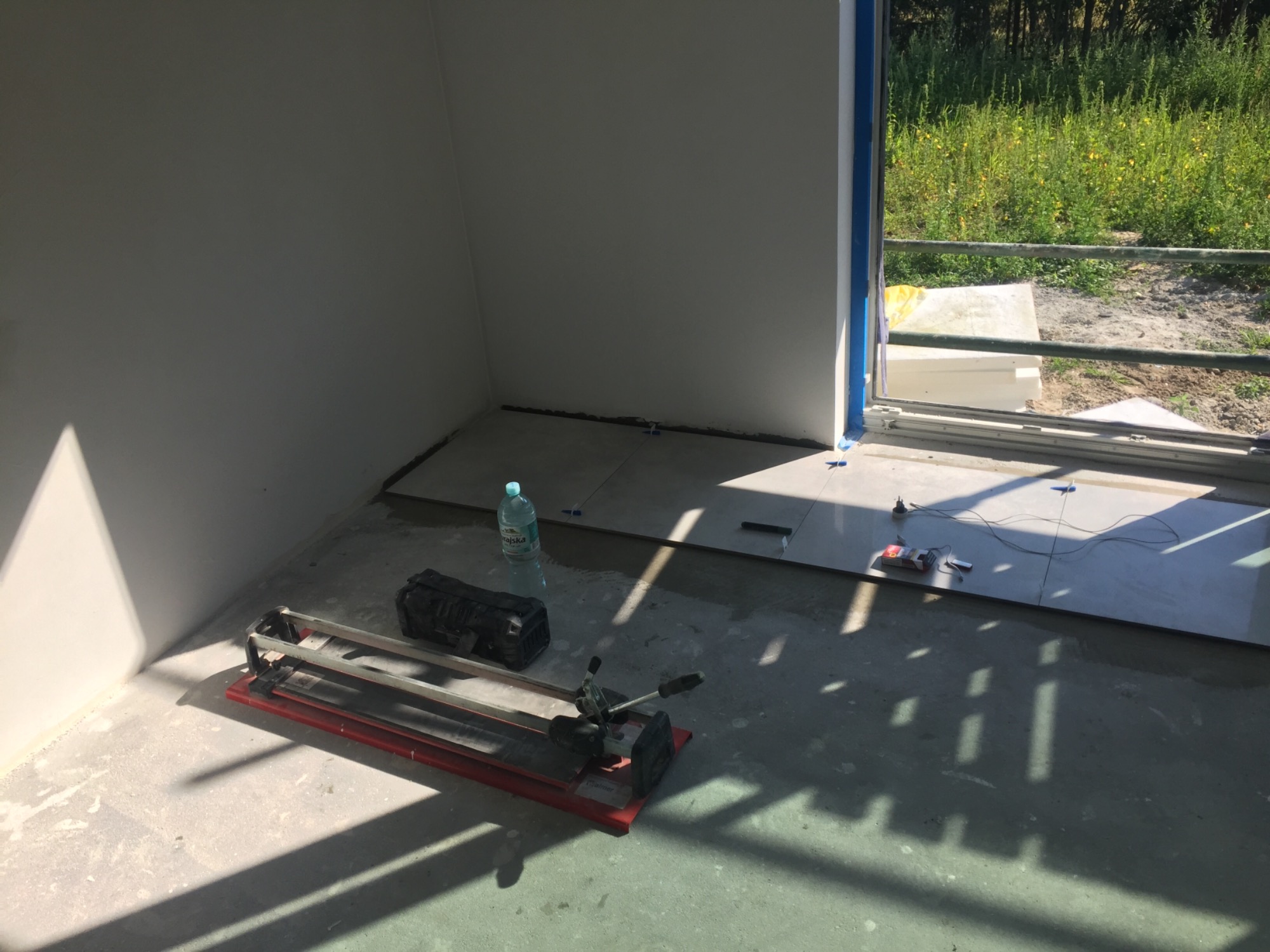 Wnętrze w trakcie układania płytek podłogowych, widoczna przecinarka do płytek, butelka wody i paczka z akcesoriami. Częściowo ułożone płytki w jasnym odcieniu szarości, widok na zewnątrz przez...