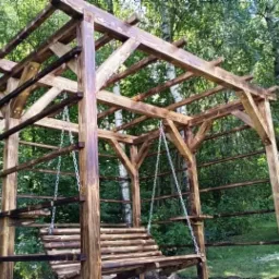 Drewniana huśtawka ogrodowa pod pergolą z surowego drewna, zawieszona na łańcuchach, w tle zielony ogród z drzewami.