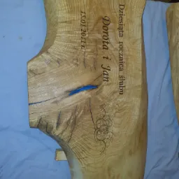 Drewniany blat z naturalną krawędzią, ozdobiony grawerem z imionami Dorota i Jan oraz datą 15.01.2011, wypełniony niebieską żywicą w pęknięciach.