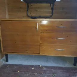 Komoda z drewna o ciepłym odcieniu, z trzema szufladami po prawej i pojedynczymi drzwiczkami po lewej, na czarnych nóżkach, ustawiona w drewnianym pomieszczeniu z widocznymi kablami powyżej.