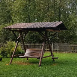 Drewniana huśtawka ogrodowa z zadaszeniem na trawniku, w tle zielone drzewa i drewniany płot. Na trawie w lewym dolnym rogu widać kurę.