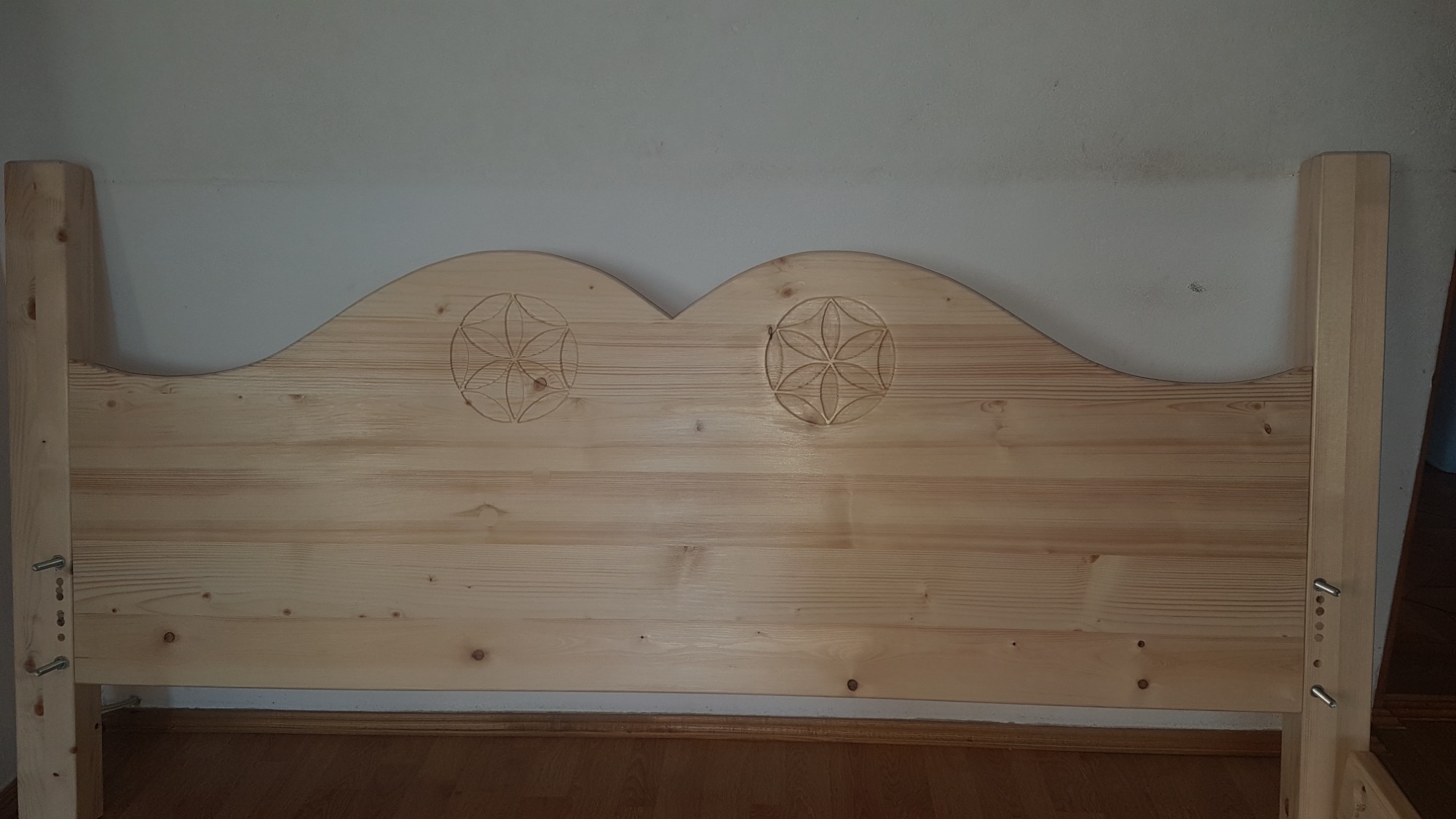 Surowe drewniane wezgłowie łóżka z wyciętymi wzorami kwiatowymi, widoczne śruby montażowe.