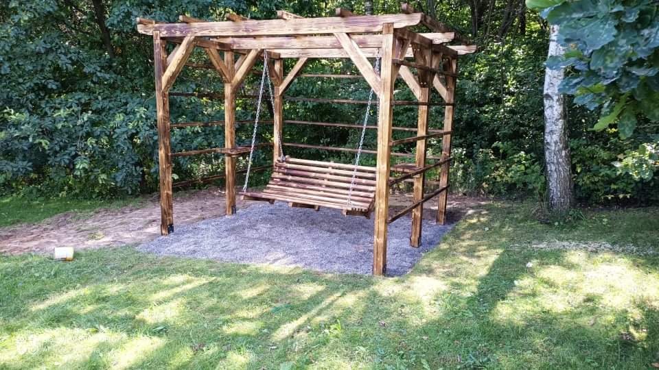 Drewniana pergola z huśtawką łańcuchową na żwirowym podłożu, otoczona zielenią drzew i trawnikiem.