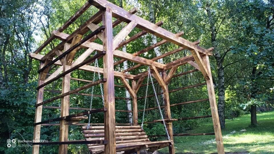 Drewniana huśtawka ogrodowa pod pergolą z surowego drewna, zawieszona na łańcuchach, w tle zielony ogród z drzewami.