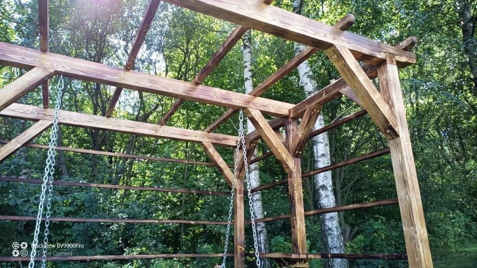 Drewniana konstrukcja pergoli z huśtawką na łańcuchach, w tle zielone drzewa.