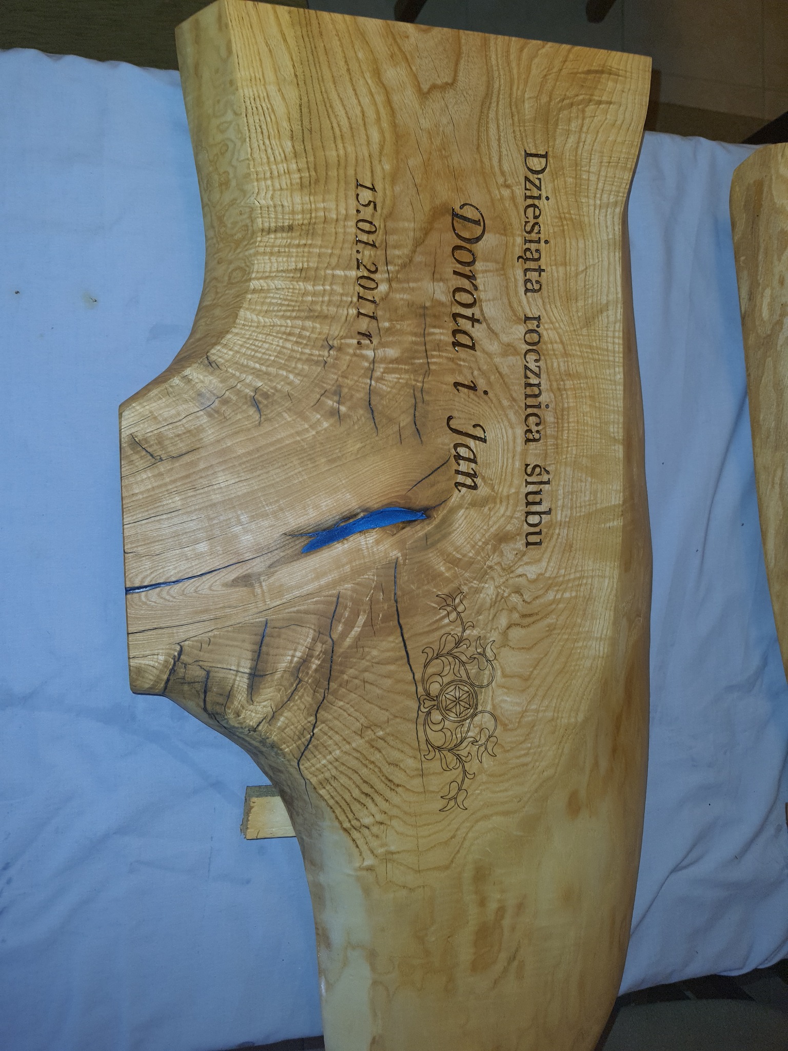 Drewniany blat z naturalną krawędzią, ozdobiony grawerem z imionami Dorota i Jan oraz datą 15.01.2011, wypełniony niebieską żywicą w pęknięciach.
