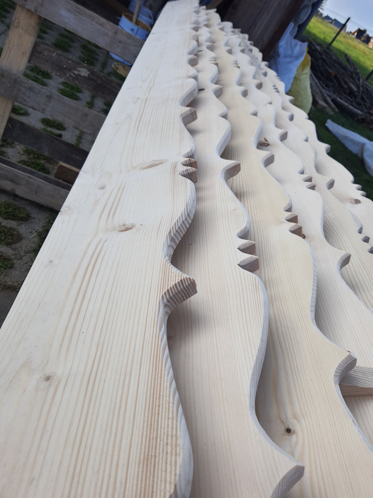 Surowe, jasne deski z wyciętymi dekoracyjnymi wzorami, ułożone jedna obok drugiej, widoczne sęki i usłojenie drewna, przygotowane do dalszej obróbki.