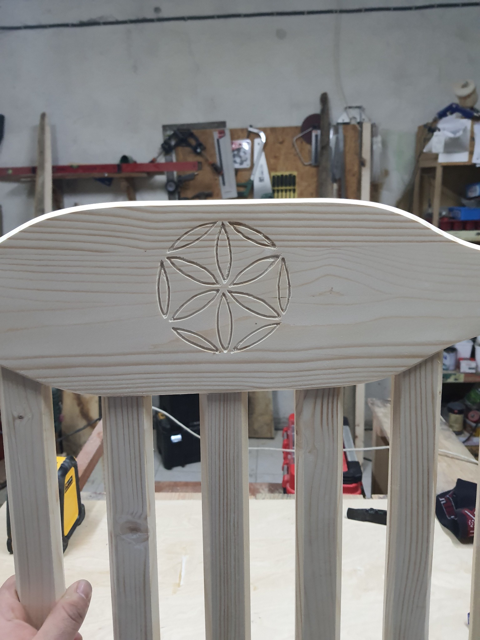 Fragment drewnianego krzesła z wyżłobionym wzorem kwiatowym na oparciu, widoczne narzędzia i warsztat w tle.