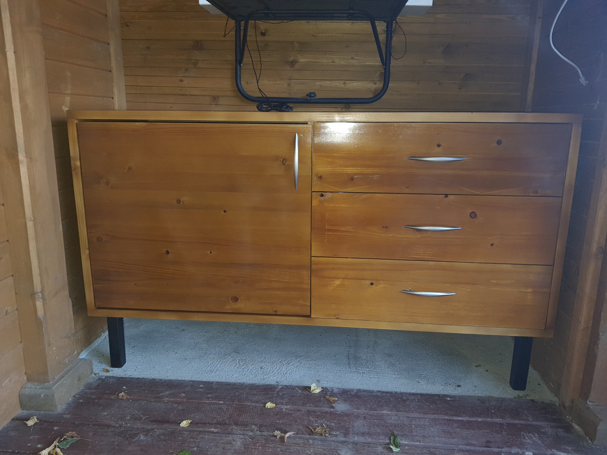 Komoda z drewna o ciepłym odcieniu, z trzema szufladami po prawej i pojedynczymi drzwiczkami po lewej, na czarnych nóżkach, ustawiona w drewnianym pomieszczeniu z widocznymi kablami powyżej.