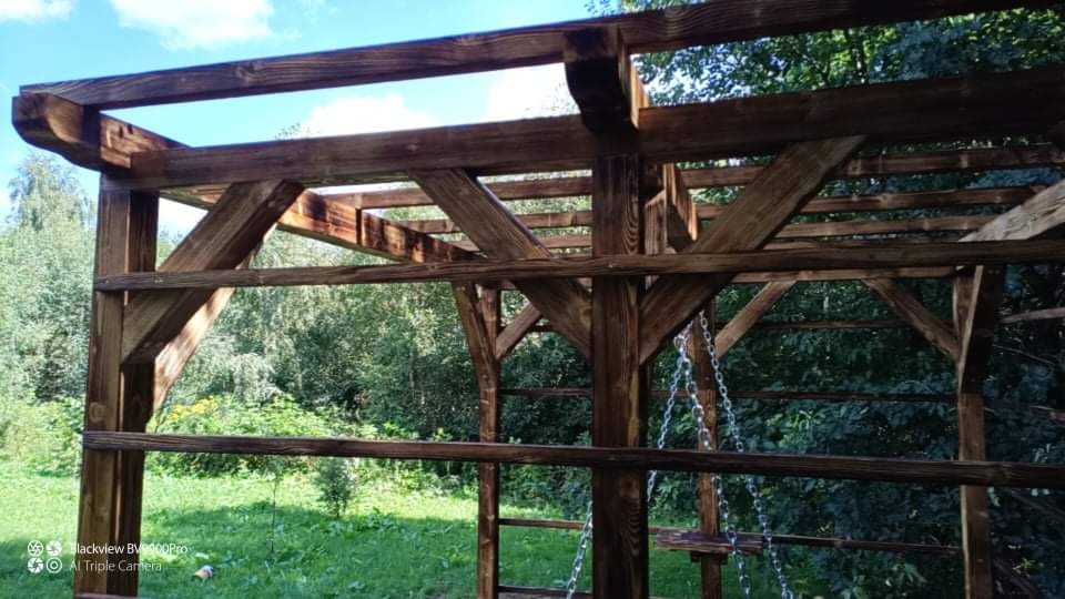 Drewniana konstrukcja z ciemnego drewna, prawdopodobnie pergola, widoczna od dołu na tle zieleni ogrodu i fragmentu nieba.