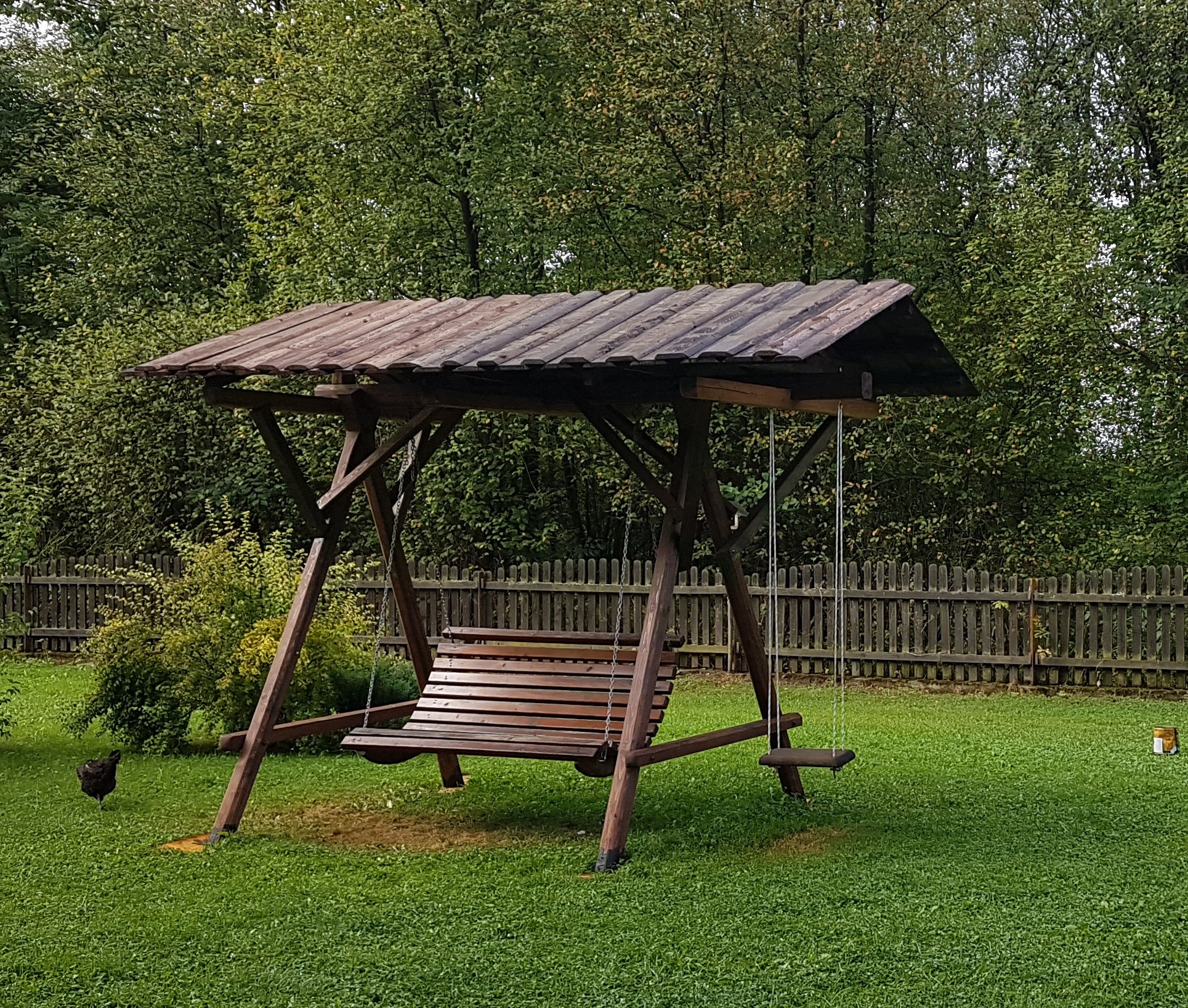 Drewniana huśtawka ogrodowa z zadaszeniem na trawniku, w tle zielone drzewa i drewniany płot. Na trawie w lewym dolnym rogu widać kurę.