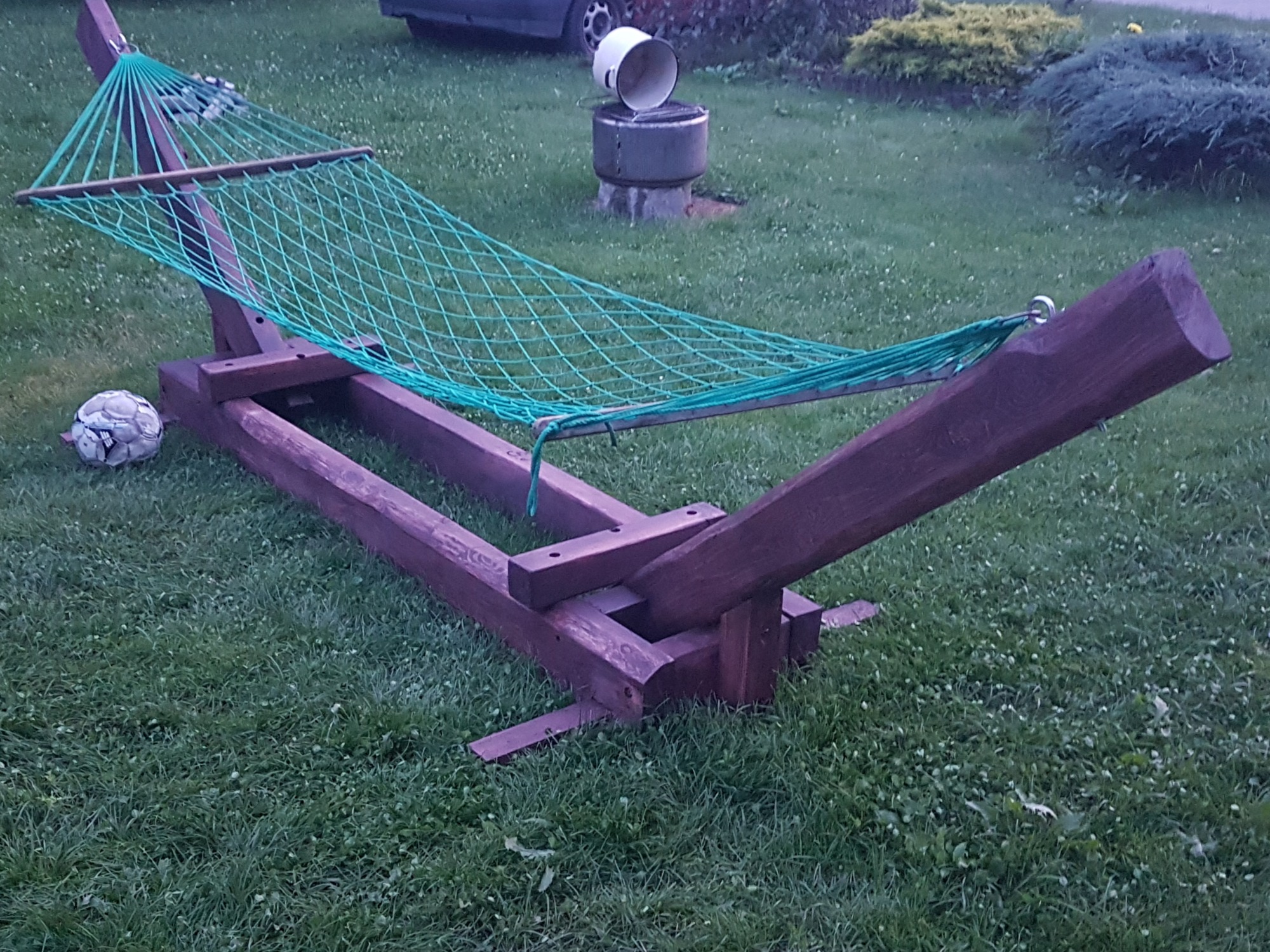 Drewniany stelaż z zieloną siatką hamaka na trawniku, obok leży piłka do nogi, w tle widoczny grill i krzewy.
