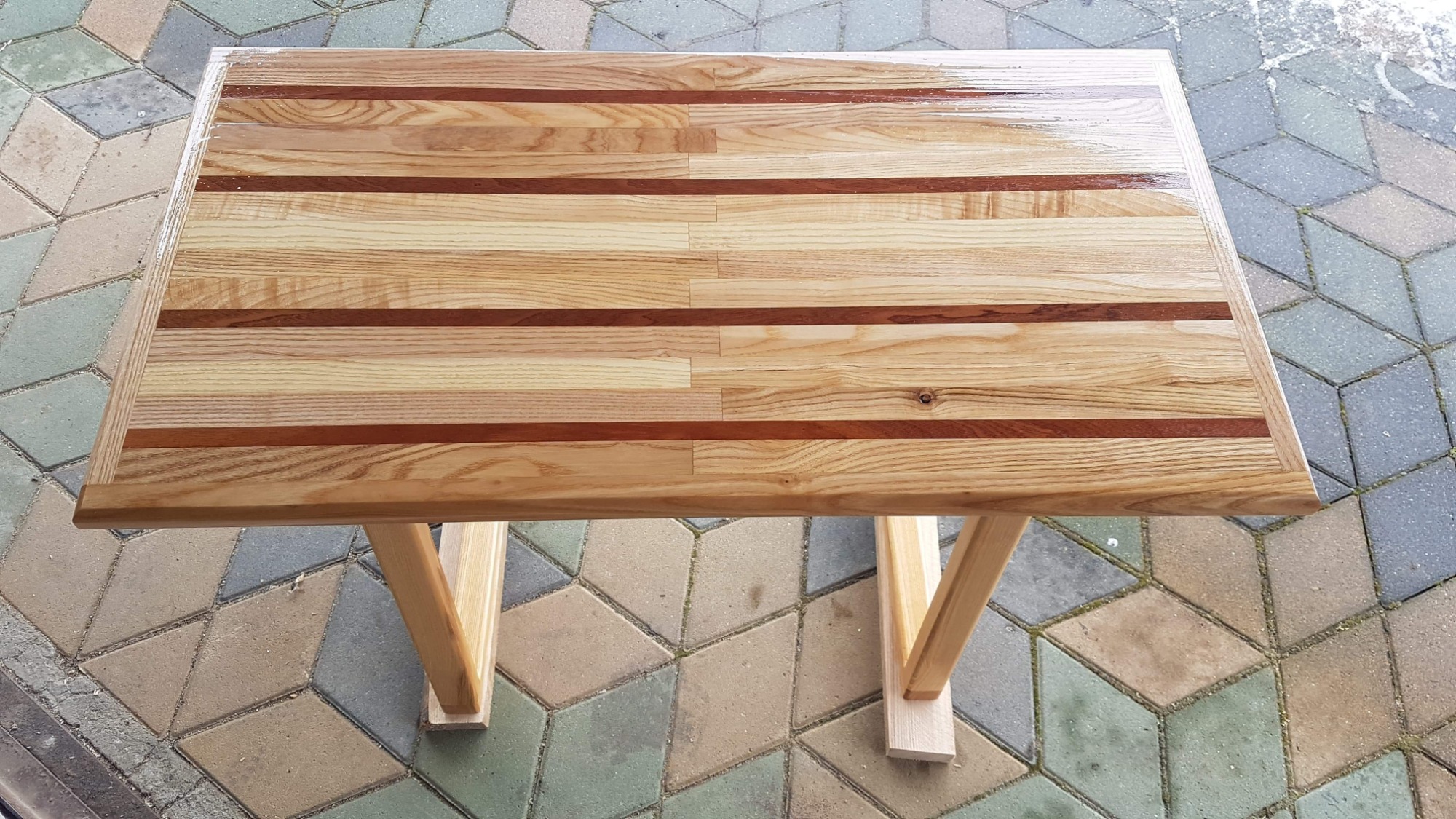 Stół z jasnego drewna z wstawkami z ciemniejszego drewna, polakierowany, z prostymi, surowymi nogami, widziany z góry na tle kostki brukowej.