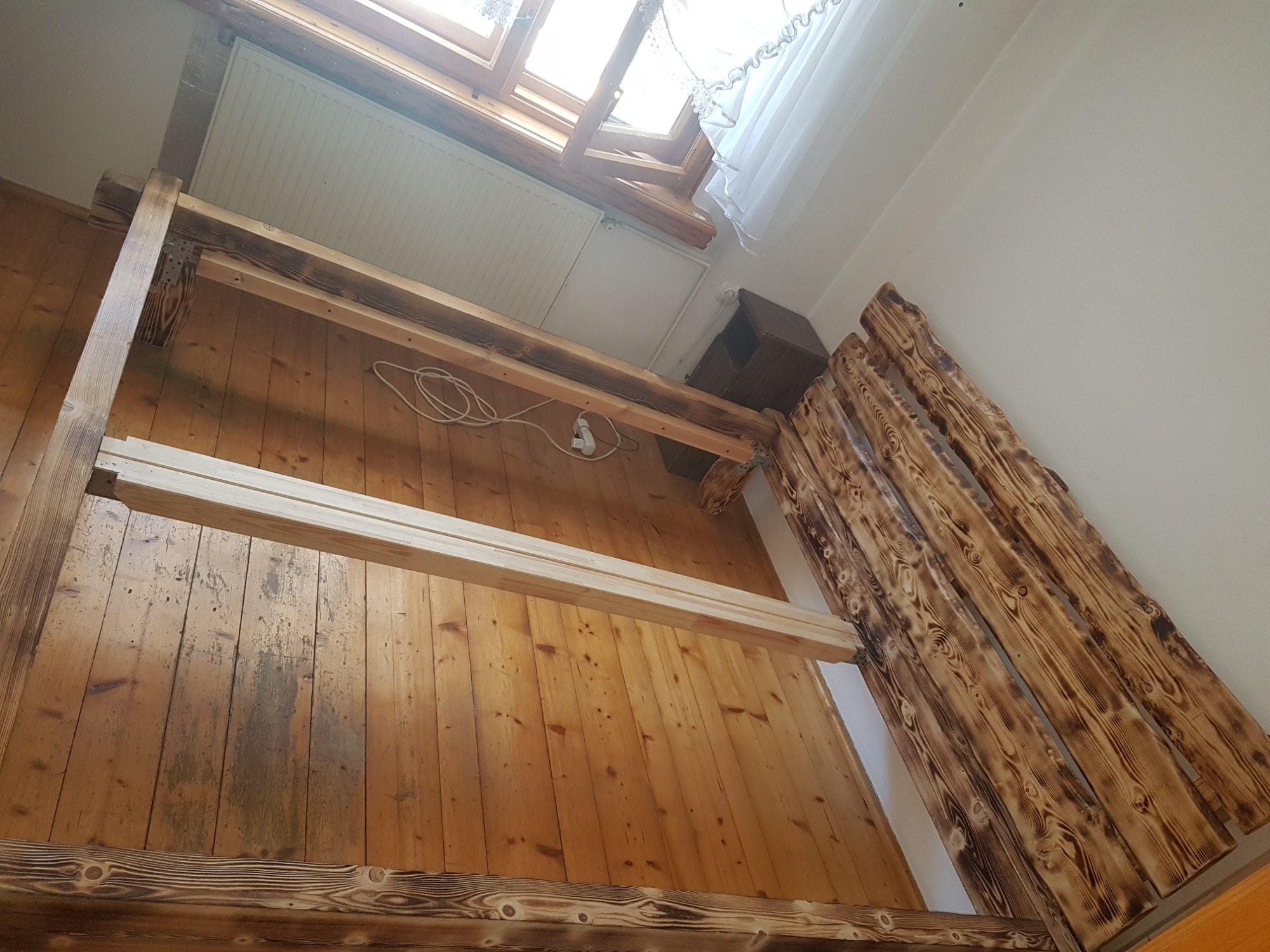 Wnętrze pokoju z drewnianą konstrukcją antresoli w trakcie budowy, widoczne deski z wypalanym wzorem, okno dachowe z białą firanką, podłoga z desek