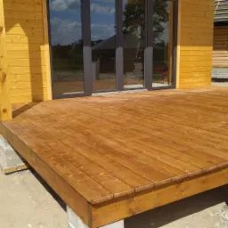 Żółta elewacja domu z drewnianą werandą wspartą na betonowych bloczkach, widoczne duże przesuwne okna z odbiciem otoczenia.