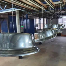 Metalowe elementy, prawdopodobnie klosze lamp, zawieszone na linkach i przygotowane do procesu obróbki powierzchniowej w hali produkcyjnej. Widoczny system transportu nad głowami i niebieska ściana...