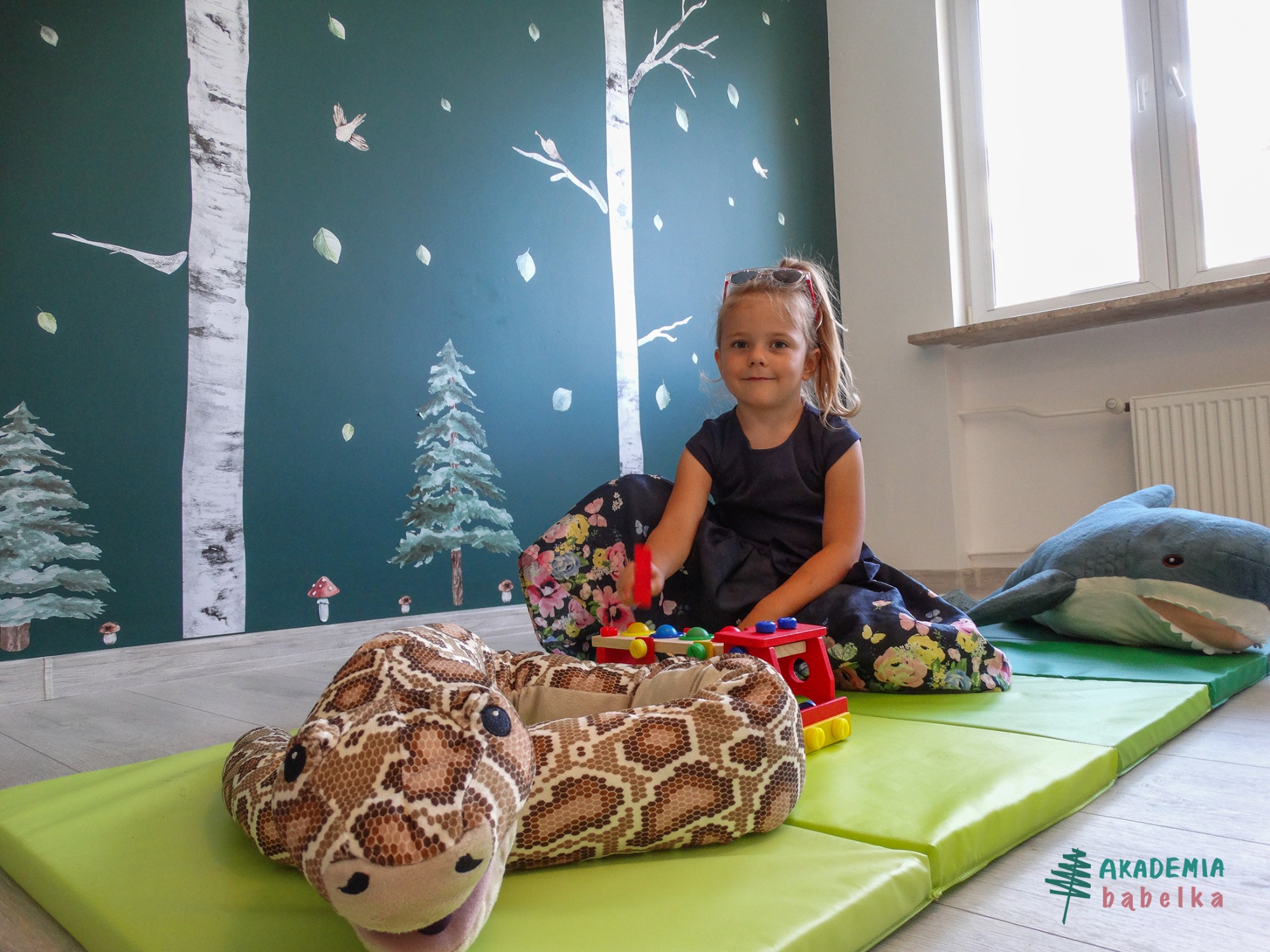 Dziewczynka w sukience siedzi na zielonej macie z zabawkami: pluszowym wężem, rekinem i drewnianą kolejką na tle ściany z motywem lasu.