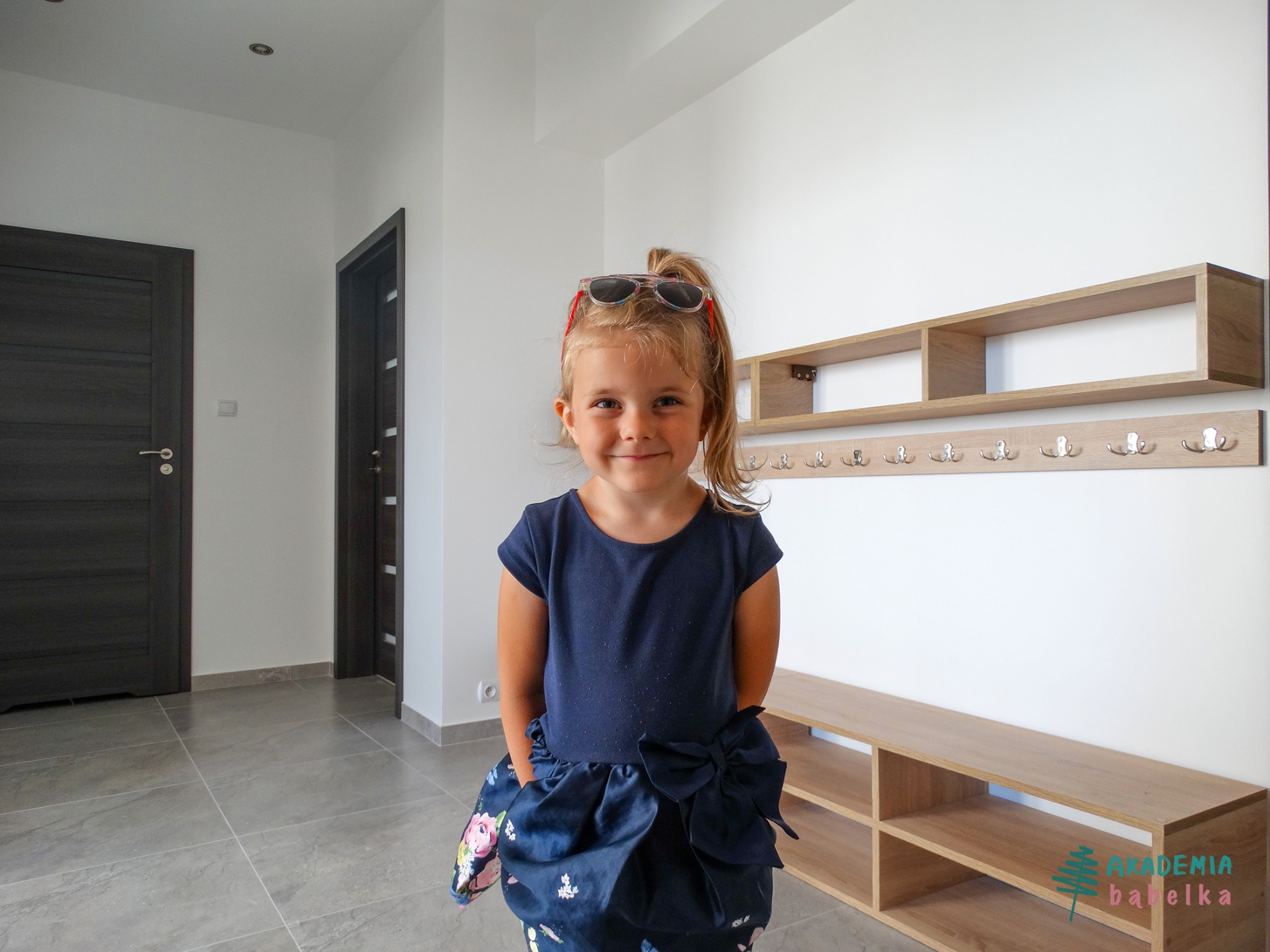 Uśmiechnięta dziewczynka w okularach na głowie, stojąca w jasnym holu z drewnianą ławką i wieszakami, widoczne drzwi i logo firmy na dole po prawej.