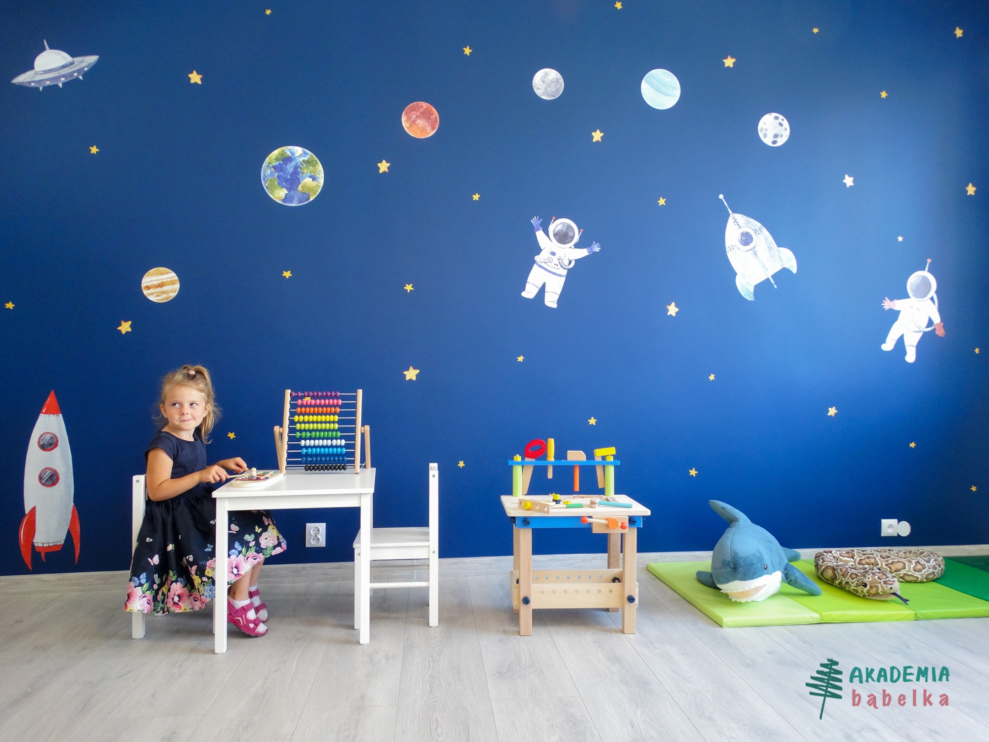 Dziewczynka siedzi przy białym stoliku z liczydłem w pokoju z motywem kosmosu na ścianie, obok stoi drewniany warsztat z narzędziami, a na podłodze leżą pluszowy rekin i wąż.