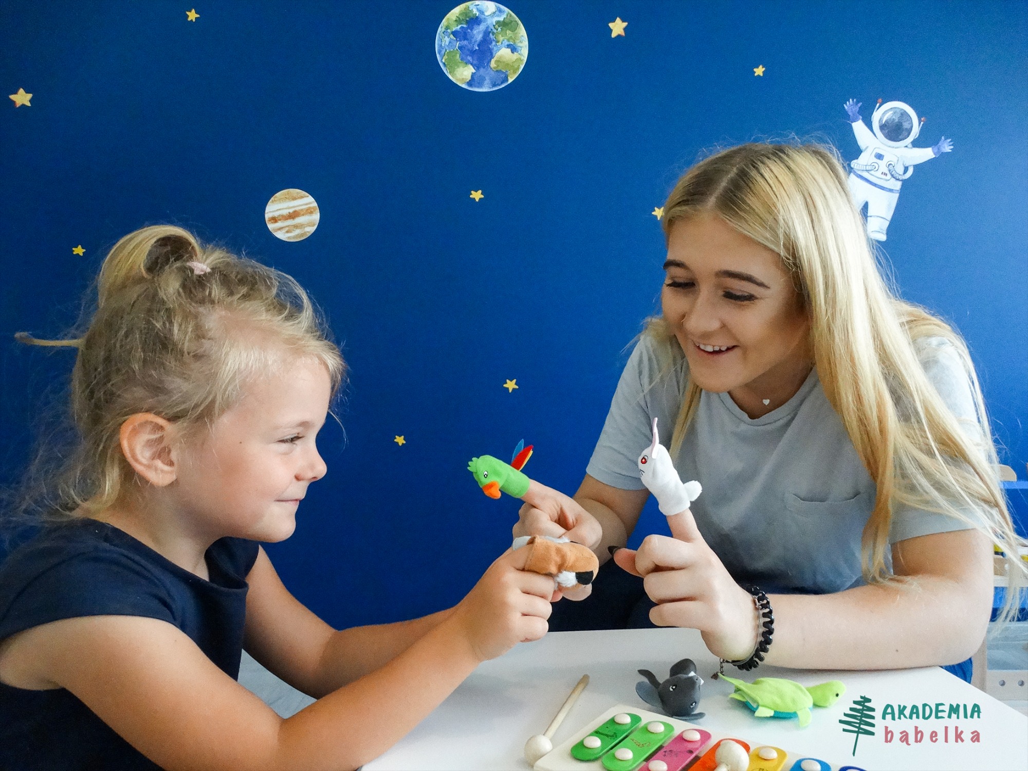 Dziewczynka i opiekunka bawią się pacynkami na palce przy stole z ksylofonem, tło ściany z motywami kosmicznymi