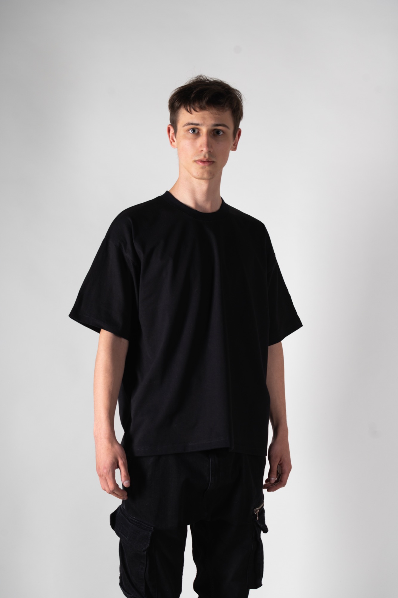 Młody mężczyzna w czarnym, luźnym T-shircie i czarnych spodniach typu cargo z kieszeniami na suwak, na jasnym tle.