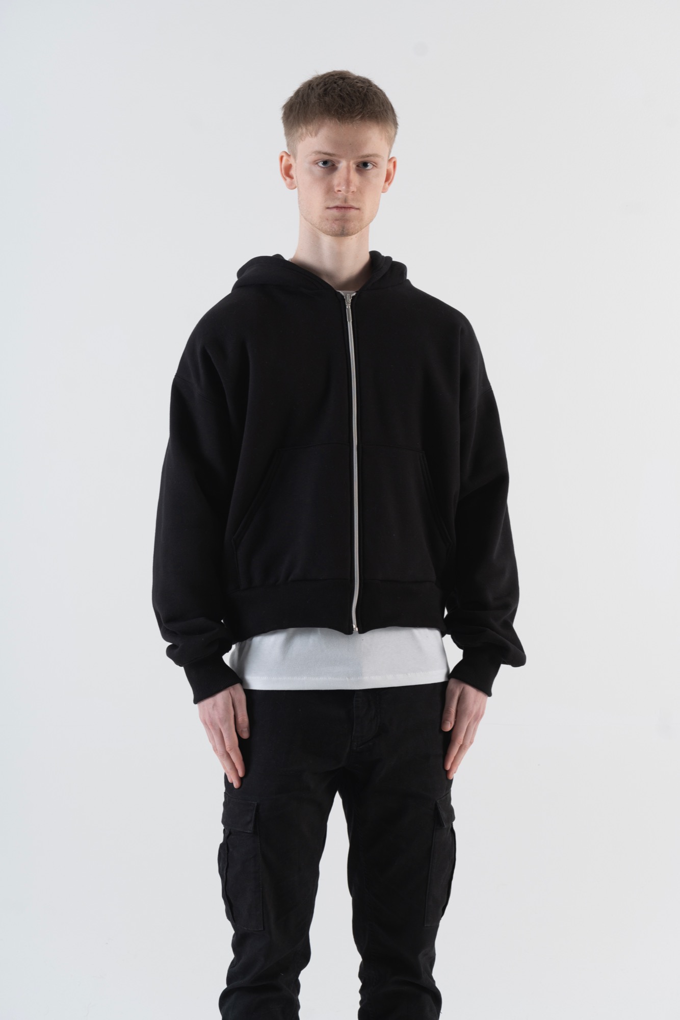 Mężczyzna w czarnej rozpinanej bluzie z kapturem, białym t-shircie i czarnych spodniach typu cargo na jednolitym białym tle.